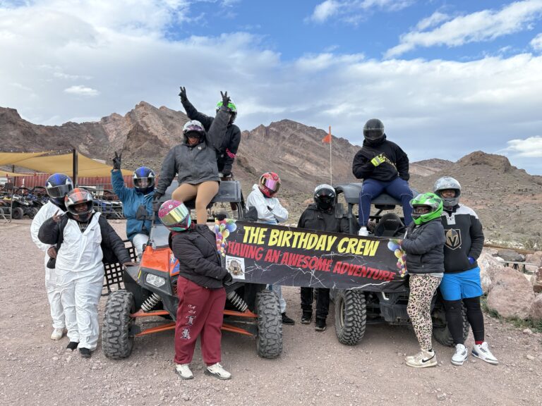 Adventurers in helmets pose by ATVs in Nevada, holding “Birthday Crew” banner with Las Vegas mountains on an epic off-road tour.