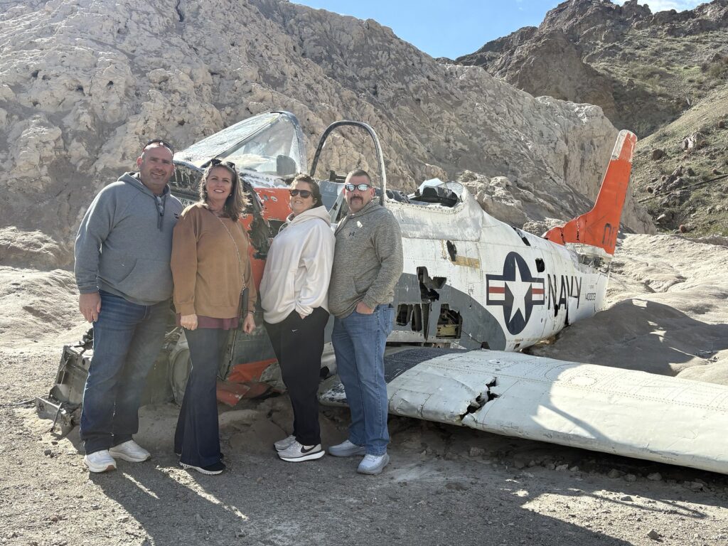 Group smiles by a wrecked US Navy plane in Nevada desert near Las Vegas, ideal for ATV tours and Ghost Town sightseeing.