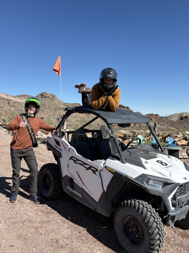 Two people pose with a Polaris RZR during an ATV tour near Las Vegas, Nevada, with desert rocks and mountains in the background.