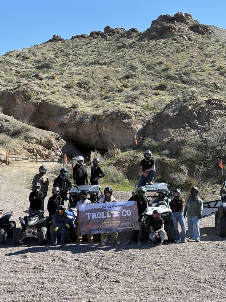 Group in helmets with ATVs and TROLLY CO banner near rocky Nevada hillside, ready for Las Vegas ATV tour adventure.
