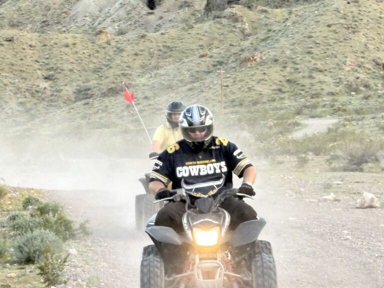 ATV tour in Nevada: Two riders on a dusty trail near Las Vegas, one in Cowboys jersey, one with red flag, exploring rocky hills.