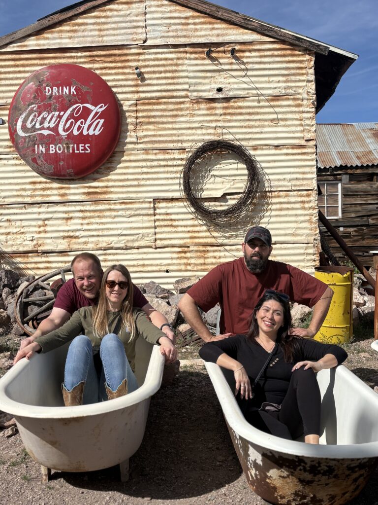 Four adults smile in old bathtubs outside a rustic Nevada ghost town with a vintage Coca-Cola sign, enjoying Colorado River sightseeing.