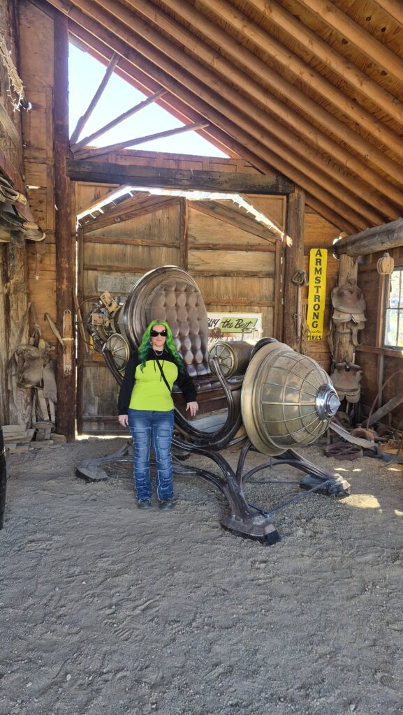 Person with green hair in sunglasses and green hoodie poses by steampunk chair in Nevada ghost town barn near Colorado River.
