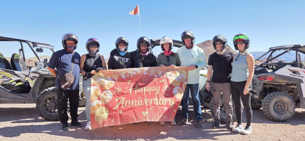 Group in helmets celebrates with a Happy Anniversary banner between two ATVs on a Las Vegas ATV tour near Nevada mountains.
