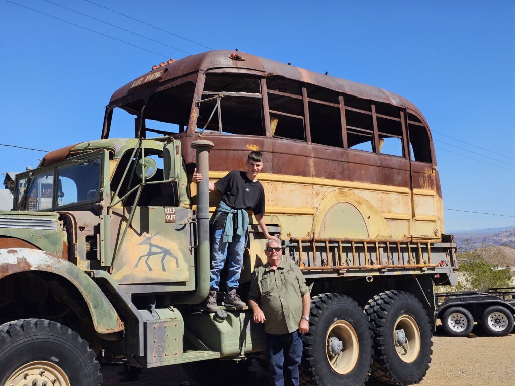 Two people pose near a rugged military vehicle with a yellow bus on top, set under Nevada's clear blue sky—perfect for Ghost Town tours.