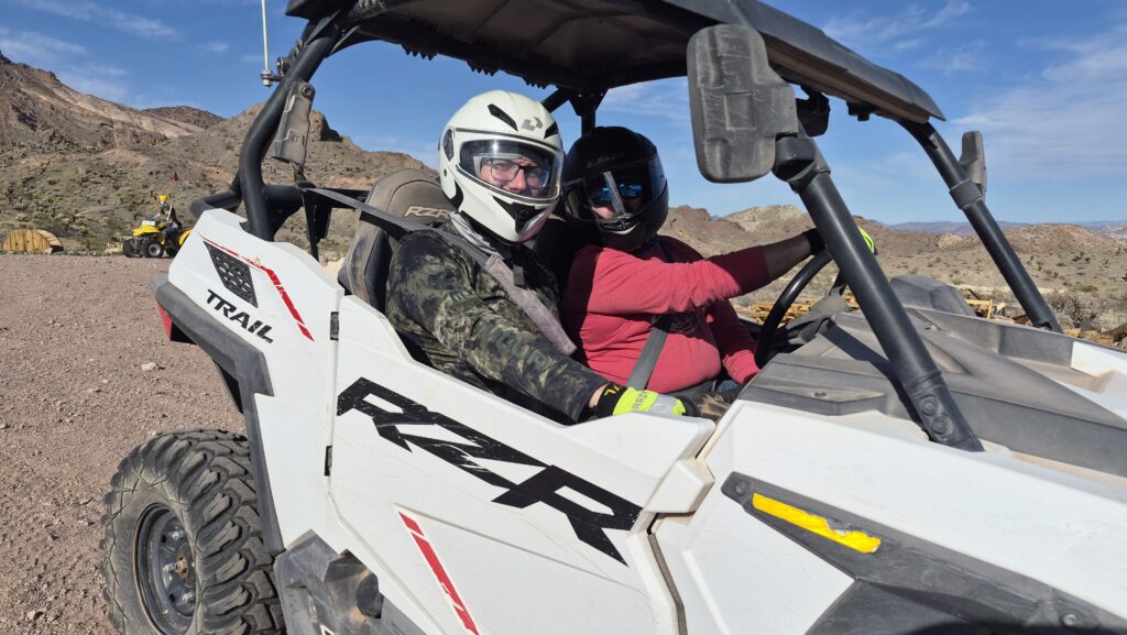 Two adventurers in helmets ride a white RZR on a rocky Nevada desert trail near Las Vegas, with ATV tours along the Colorado River.
