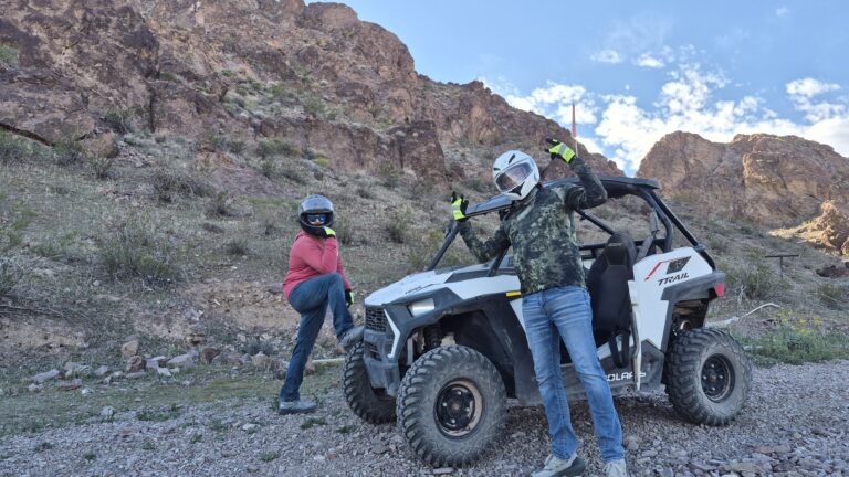 Two people in helmets pose by a white RZR on a Nevada desert ATV tour near Las Vegas, rocky hills and blue sky behind.