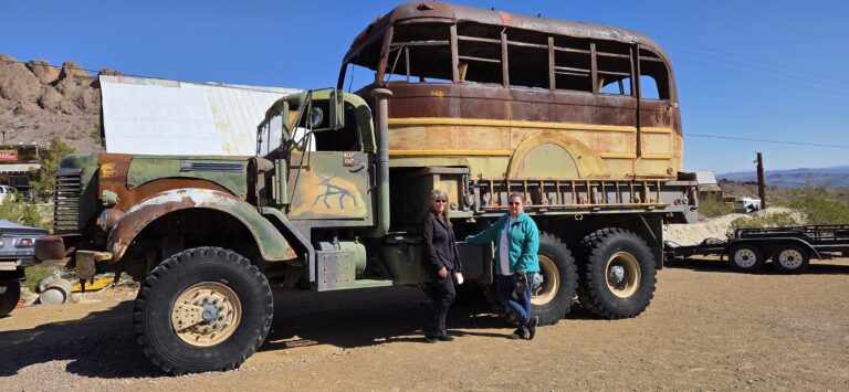 Two people by a rusted camo truck with bus top in Nevada, near rocky hills—perfect for Ghost Town sightseeing or ATV tours.