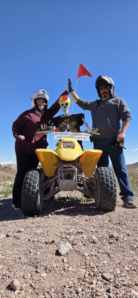 Riders high-five by a yellow ATV with a skeleton in sunglasses near Las Vegas on rocky terrain during an off-road tour.