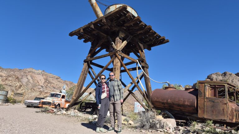 Smiling duo under a wooden water tower in a Nevada ghost town, old vehicles around—perfect for Las Vegas ATV or RZR tours.