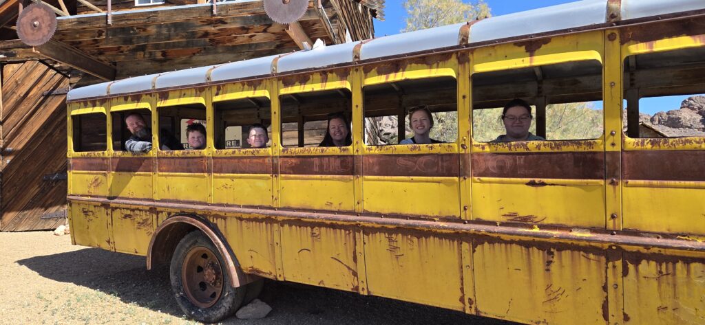 Seven smiling people look out an old yellow school bus near a wooden building on a sunny Nevada ghost town sightseeing tour.