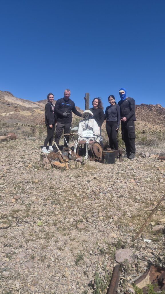 Four people in black gear stand near a white robot figure in Nevada’s rocky desert near Las Vegas, mountains and blue sky behind.