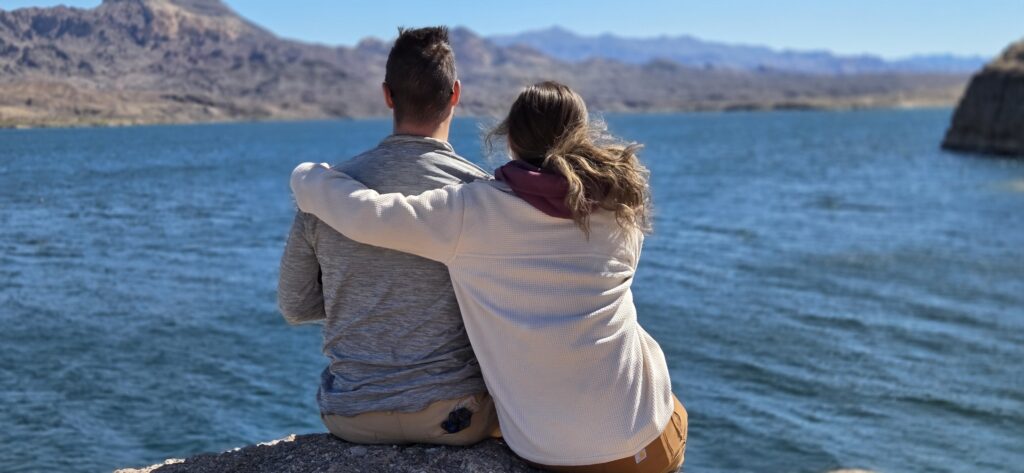 Couple sits on a rock by the Colorado River near Las Vegas, Nevada, enjoying water views and distant mountains under a clear sky.