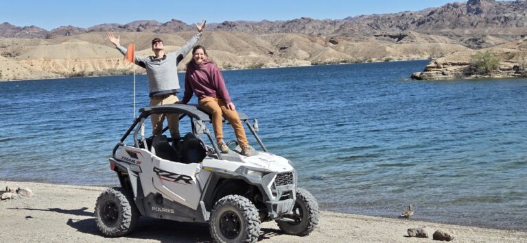 Two people enjoy a joyful RZR off-road ride by the Colorado River near Las Vegas, Nevada under a clear blue sky.