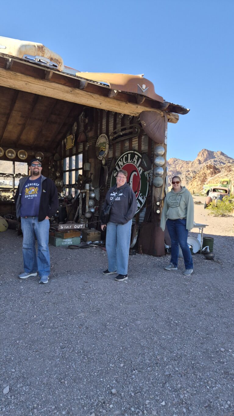 Three people at a Nevada ghost town shed with vintage signs, mountains, and old car—perfect for Las Vegas ATV tours or sightseeing.
