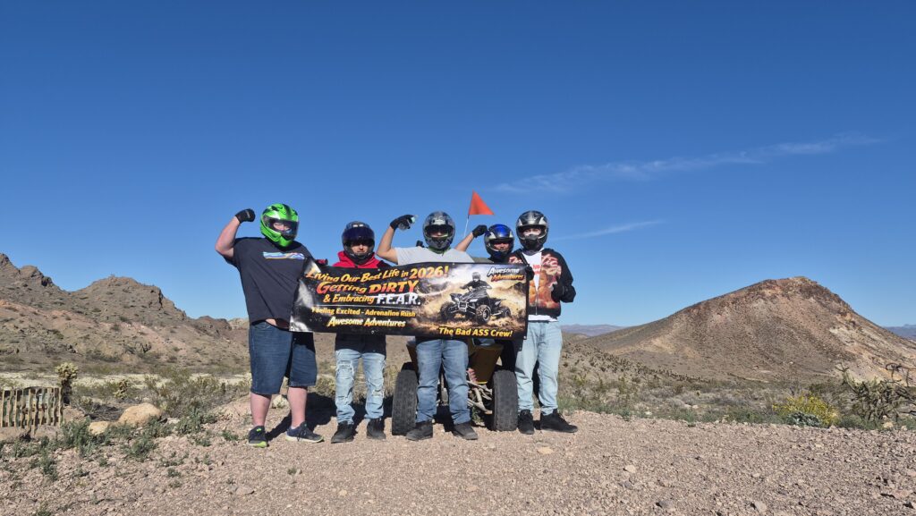 Group in helmets and outdoor gear celebrates on Nevada rocky terrain near Las Vegas, holding a banner after an ATV desert tour.