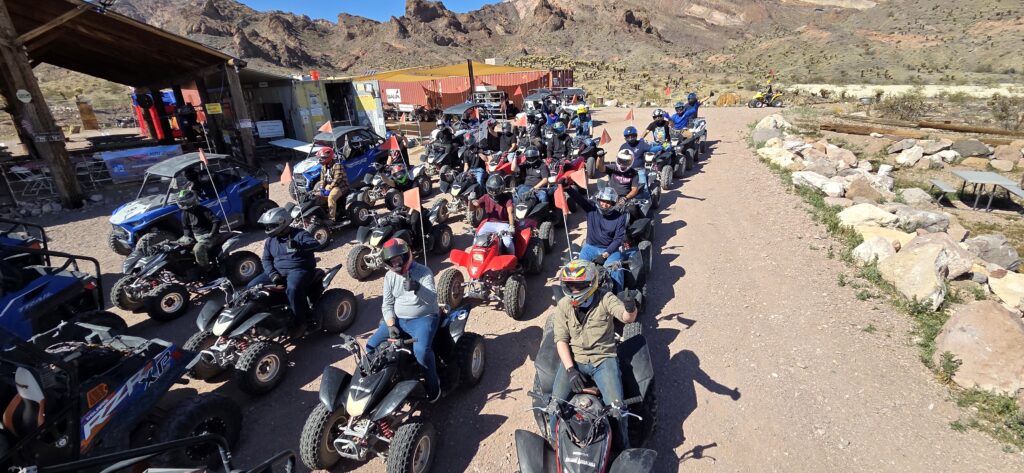 Group wearing helmets on ATVs preps for a Nevada desert RZR tour near rocky hills and cabins under Las Vegas’ clear blue sky.