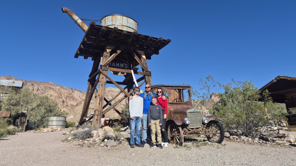 Three visitors stand by a rusted car and water tower in a Nevada desert ghost town near Las Vegas, perfect for sightseeing tours.