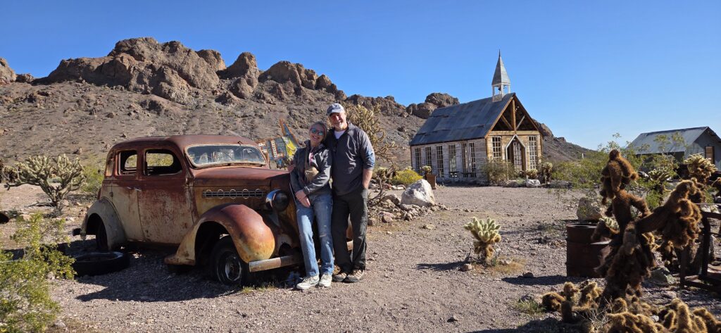 A smiling couple by a rusty old car in Nevada desert near Las Vegas ghost town, rocky hills, and rustic church under blue sky.