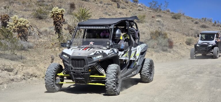 RZR off-road vehicles cruise a dusty Nevada desert trail near Las Vegas with helmeted riders, cacti, and clear blue skies.