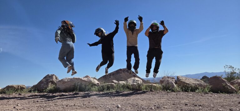 Four friends in helmets leap joyfully on a Nevada desert ATV tour near Las Vegas, blue sky and rocky landscape behind them.