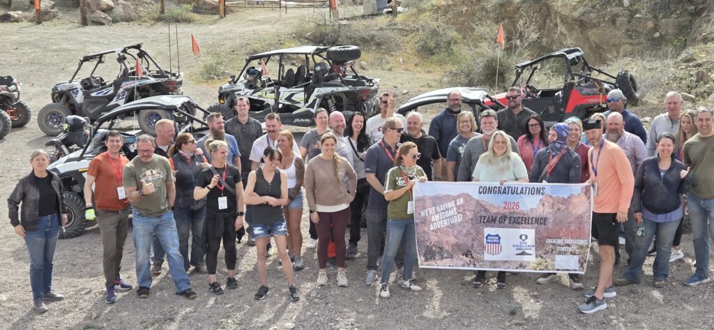 Happy group in Nevada poses by off-road vehicles after RZR tour, holding “Congratulations 2023 Team of Excellence” banner.