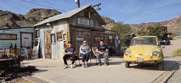 In a Nevada ghost town near Las Vegas, three relax by a vintage shop, old yellow car, desert views & mountain backdrop.