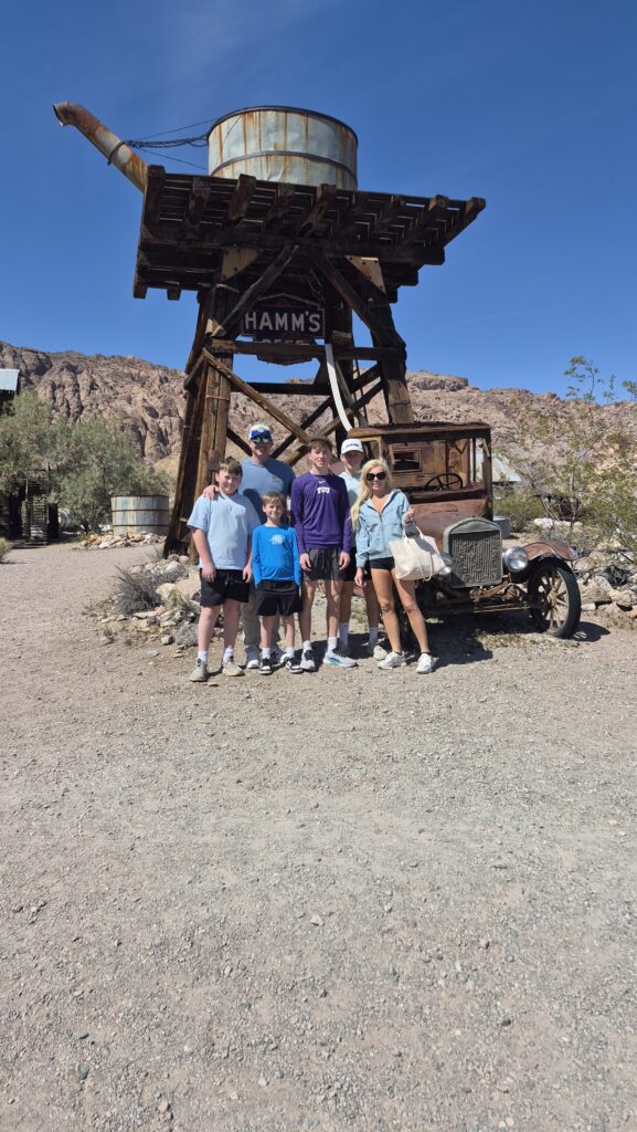 Family of five at Nevada ghost town near Las Vegas, posing by HAMMS water tower and old car amid desert hills under blue sky.