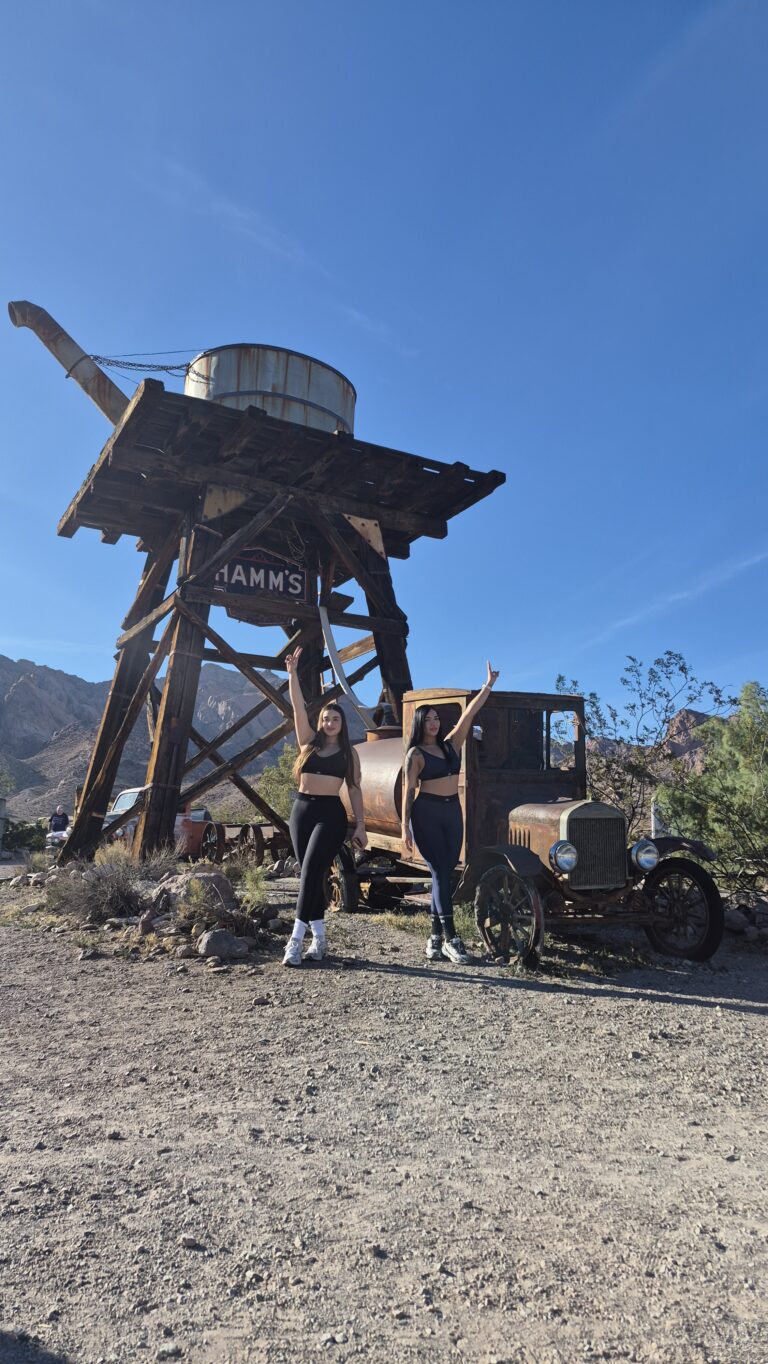Two women pose by a vintage car and wooden water tower in a Nevada desert ghost town near Las Vegas, perfect for ATV tours.