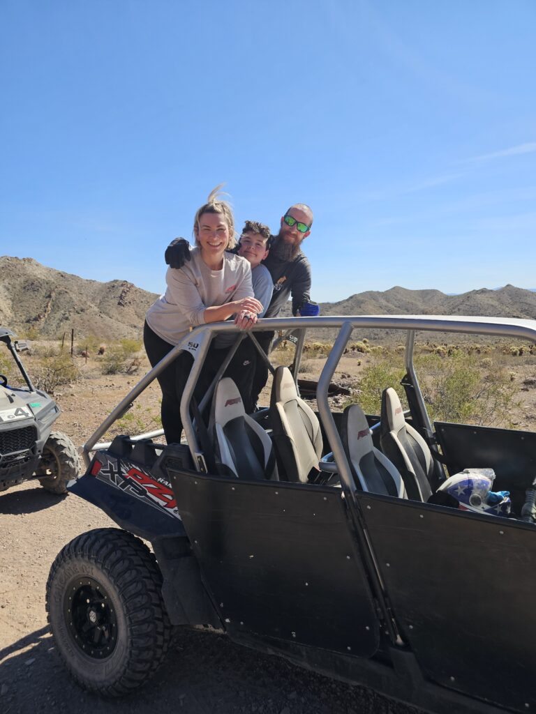 Smiling friends pose by an off-road vehicle in Nevada desert near Las Vegas, with distant ATVs and mountains under blue sky.