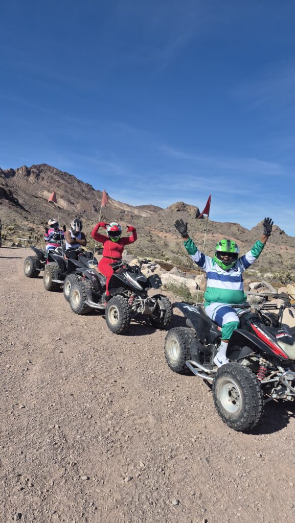 ATV tour group in colorful gear celebrates on desert trail near Las Vegas, Nevada, with mountains and blue sky in the background.