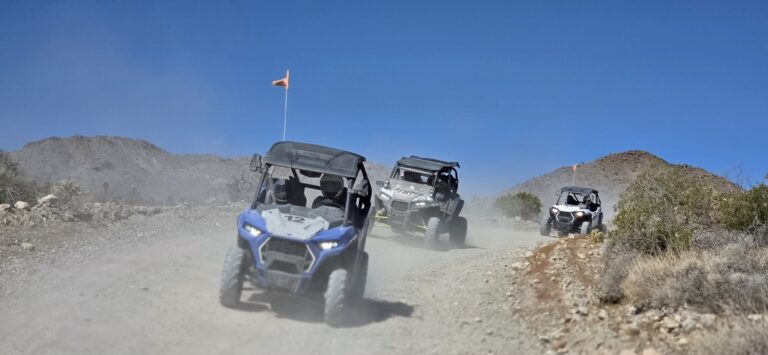 RZR off-road vehicles kick up dust on a Nevada desert trail near Las Vegas, orange flags flying, with mountains in the distance.