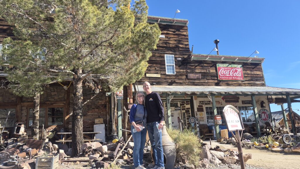 Smiling pair in front of a rustic Nevada ghost town building, Coca-Cola sign, desert plants—perfect for Las Vegas sightseeing.