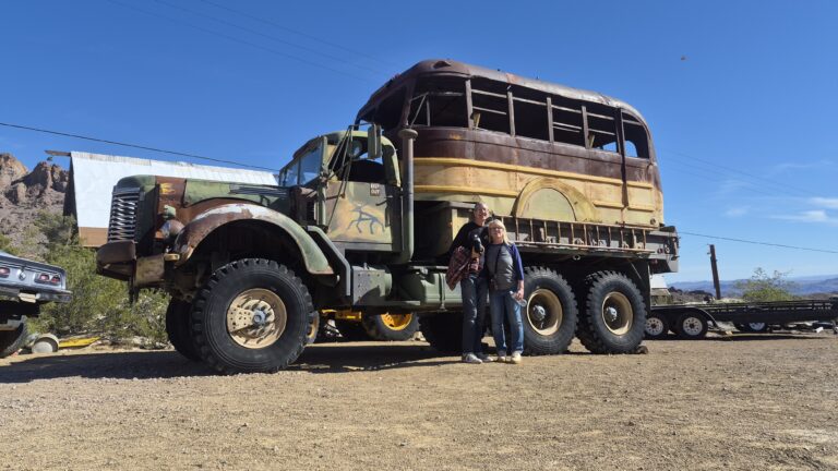 Two people by an old military truck and rusted bus in a Nevada desert lot near Las Vegas, perfect for Ghost Town sightseeing.