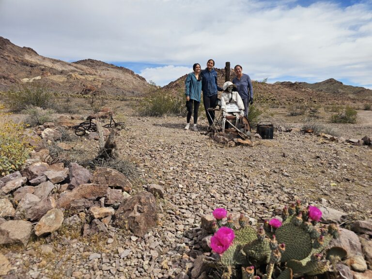 Group poses by metal statue in Nevada desert with blooming cactus, mountains, near Las Vegas—ideal for Ghost Town sightseeing.