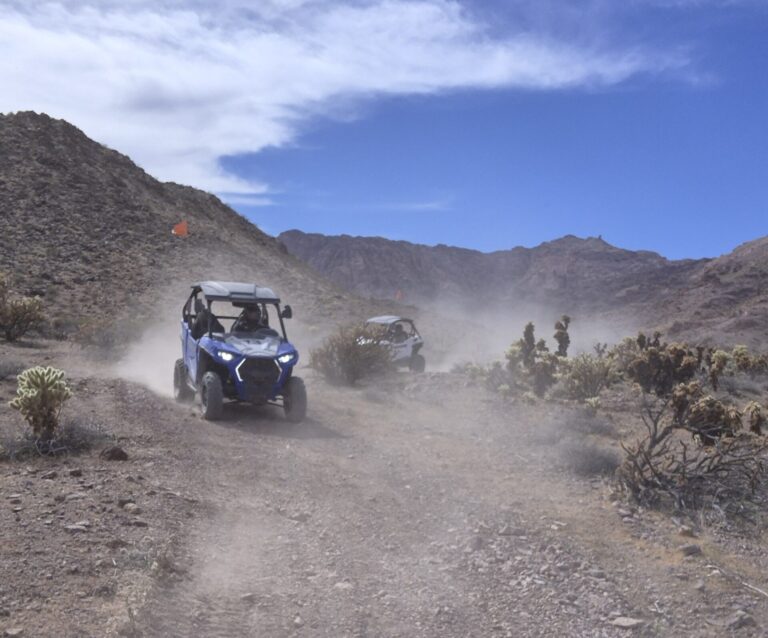 ATVs explore a dusty Nevada desert trail near Las Vegas, with rocky hills, sparse bushes, and scenic blue skies in the background.