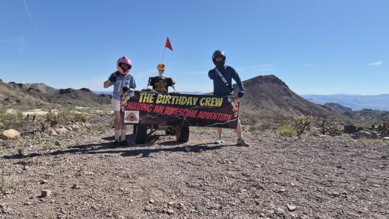 Three riders in racing gear by an ATV near the Colorado River, Nevada, with a "Birthday Crew" adventure banner—ATV tour fun!.