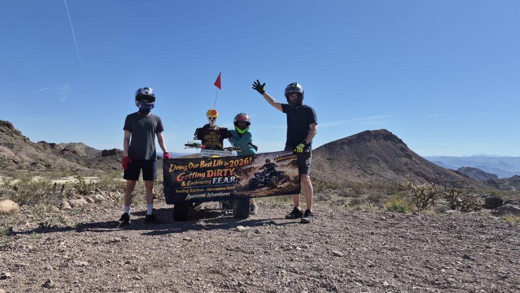 Three adventurers in helmets hold a banner on Nevada rocks near Las Vegas, with desert hills, blue sky—perfect for ATV or RZR tours.