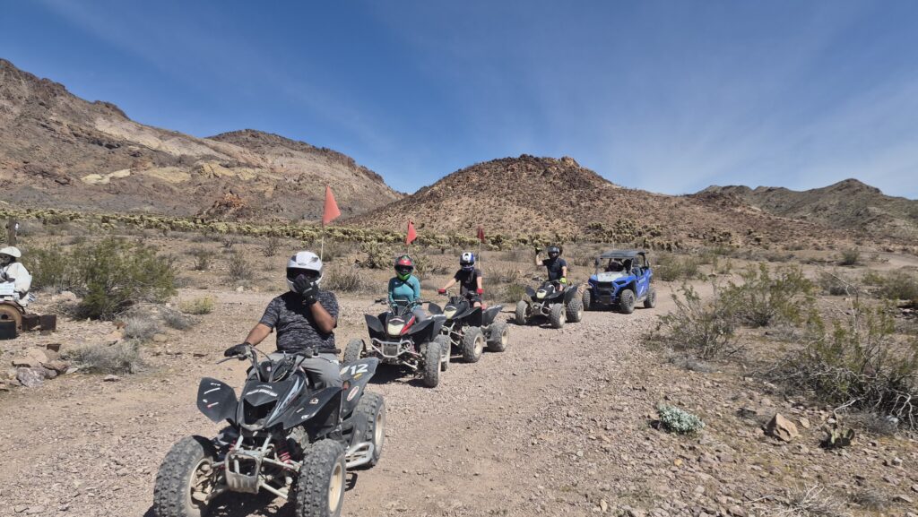 Adventurers in helmets ride ATVs and a blue UTV on a rocky Nevada desert trail near Las Vegas with mountains in the background.