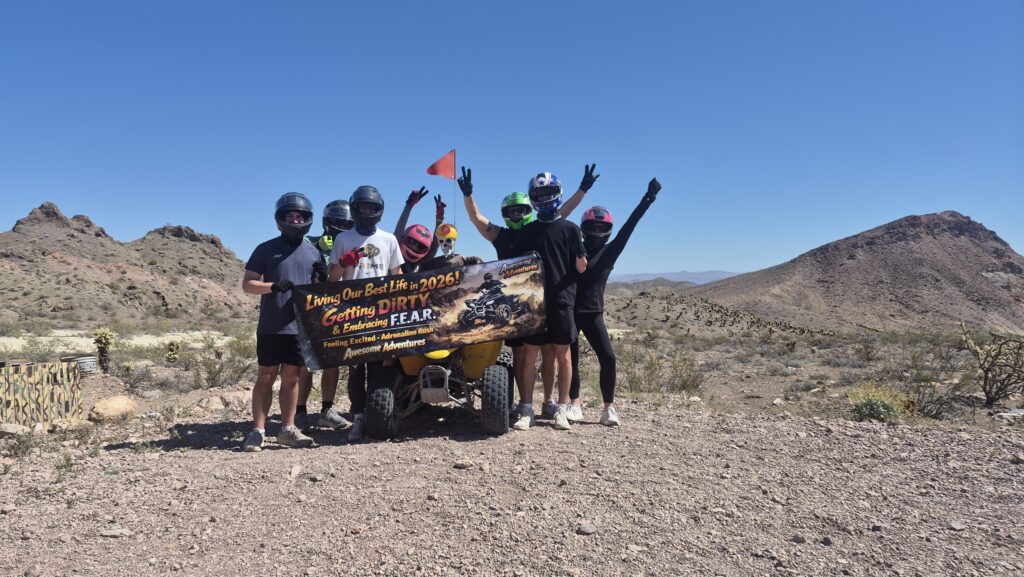Six friends in helmets pose by an ATV with a “Living Our Best Life—2026!” banner on a Nevada desert ATV tour near Las Vegas.