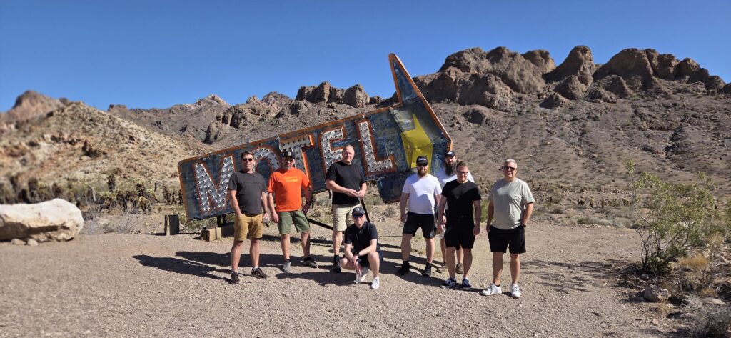 Group of men by vintage arrow-shaped MOTEL sign near Nevada ghost town, desert backdrop, perfect for Las Vegas ATV or RZR adventures.