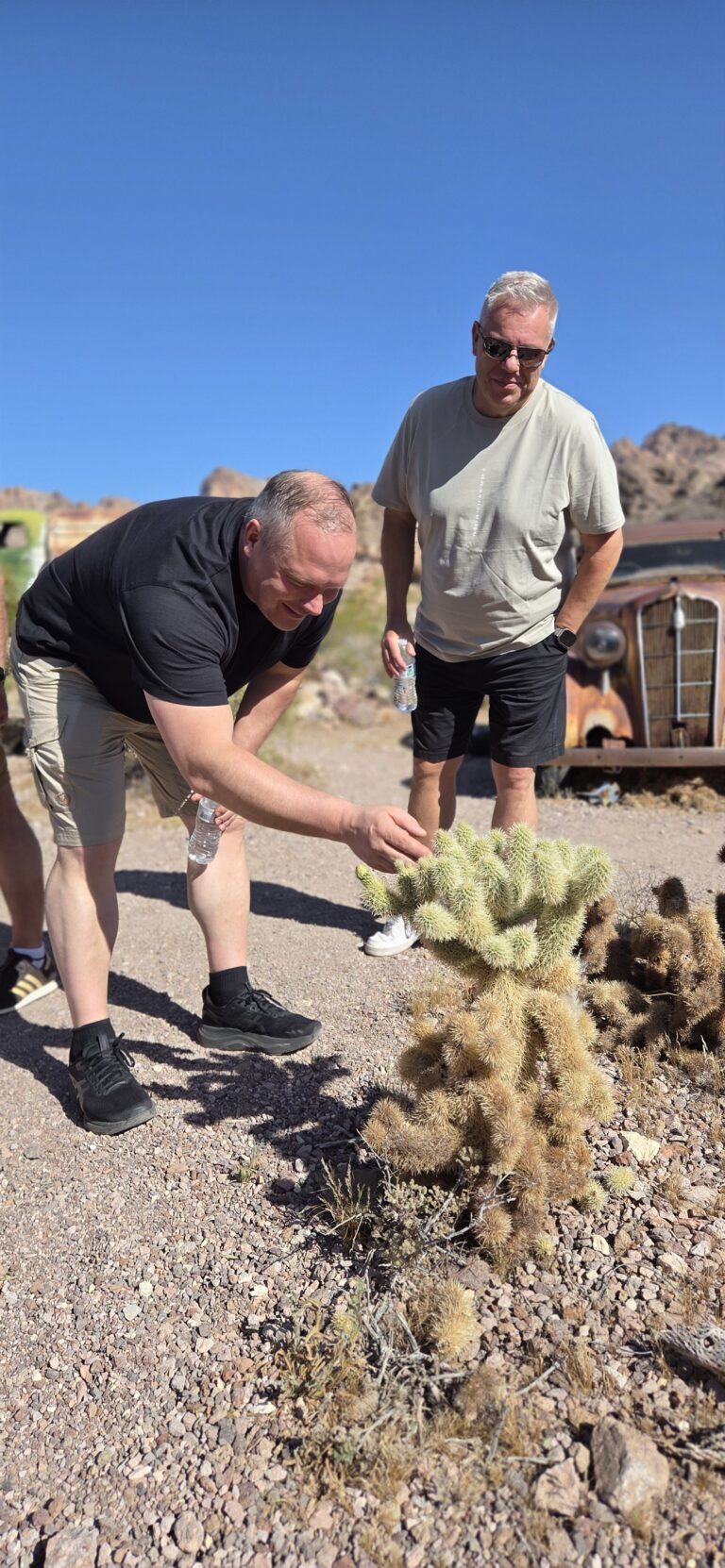 Two men explore a Nevada ghost town near the Colorado River, with old cars, rocky hills, and cacti under the Las Vegas sun.