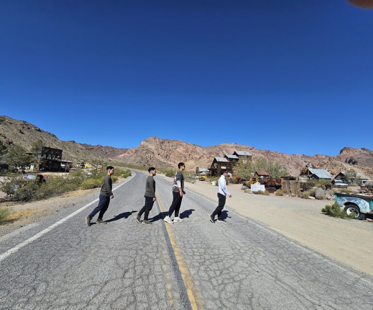 Four people walk in a line on a Nevada ghost town road near the Colorado River, mimicking Abbey Road with mountains behind.
