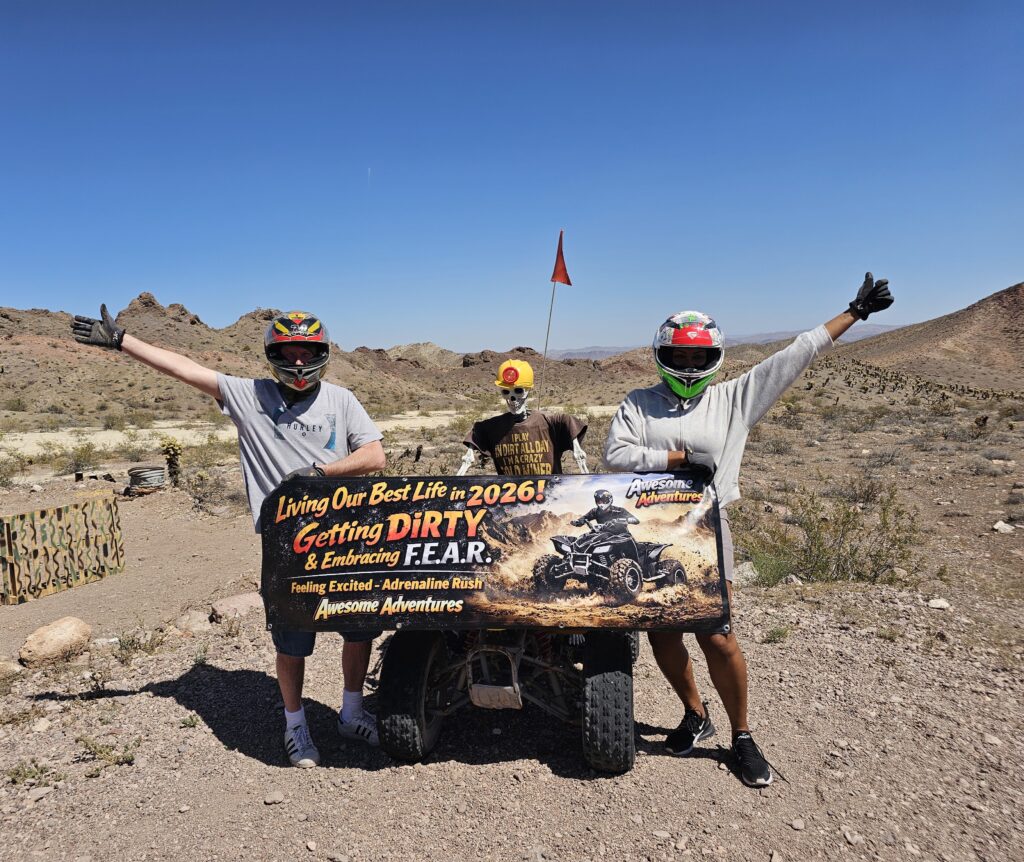 Three in helmets pose in Nevada desert near Las Vegas, holding a “Living Our Best Life 2026” banner during an ATV adventure.