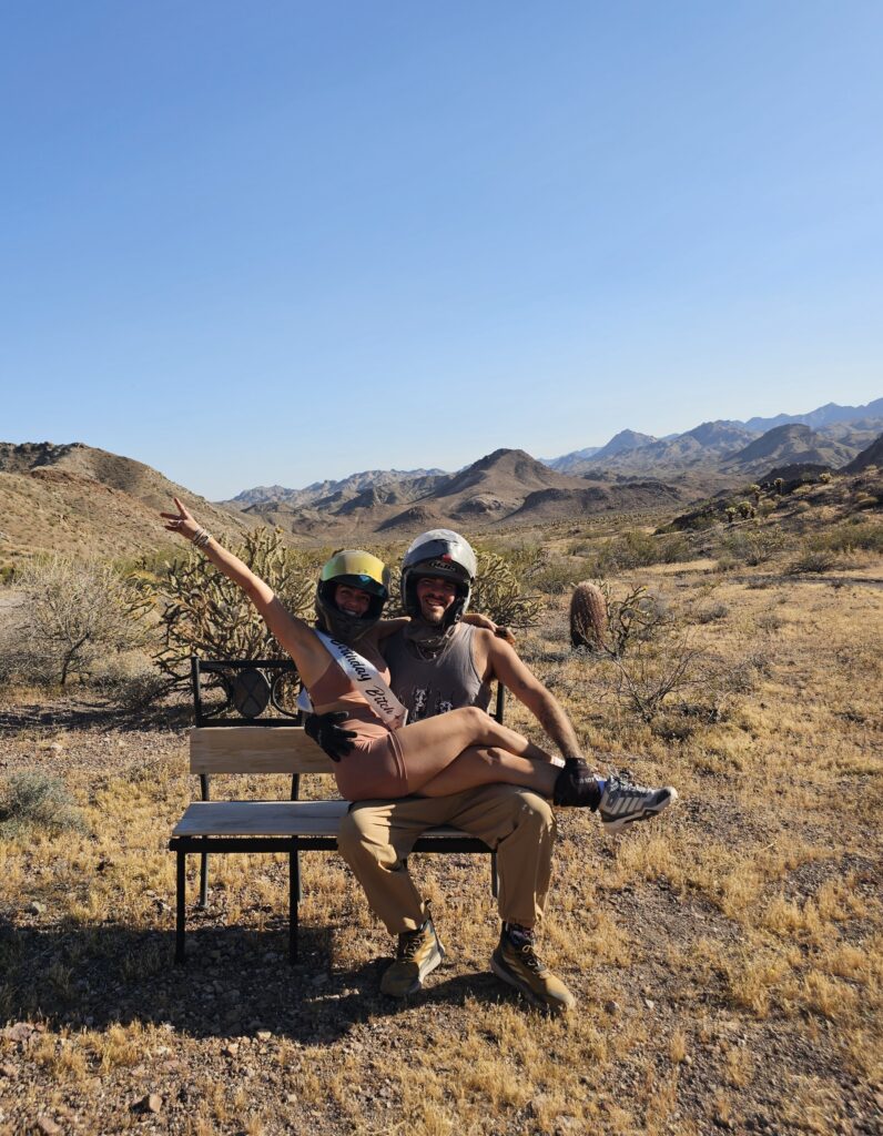 Two people in helmets relax on a desert bench near Las Vegas after an ATV tour, with Nevada mountains and clear blue sky behind.