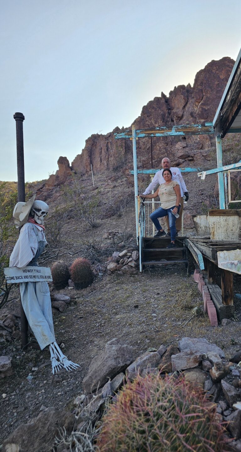 Smiling couple on wooden steps near Nevada desert; cowboy skeleton greets near cacti at Las Vegas Ghost Town sightseeing spot.