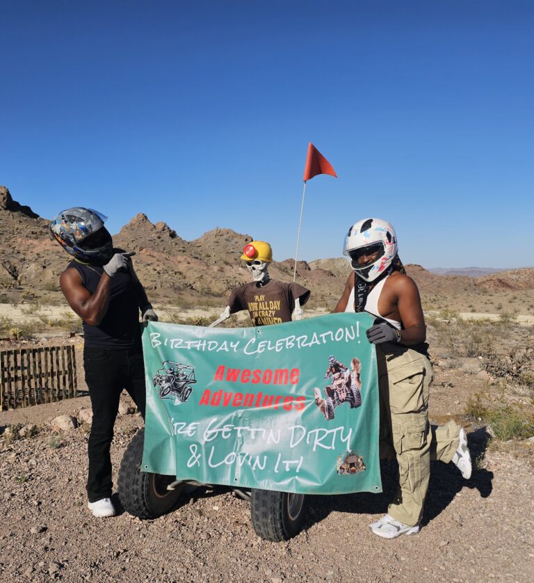 Three helmeted friends in Nevada desert hold a “Birthday Celebration” banner; skeleton joins ATV tour near Colorado River, Las Vegas.