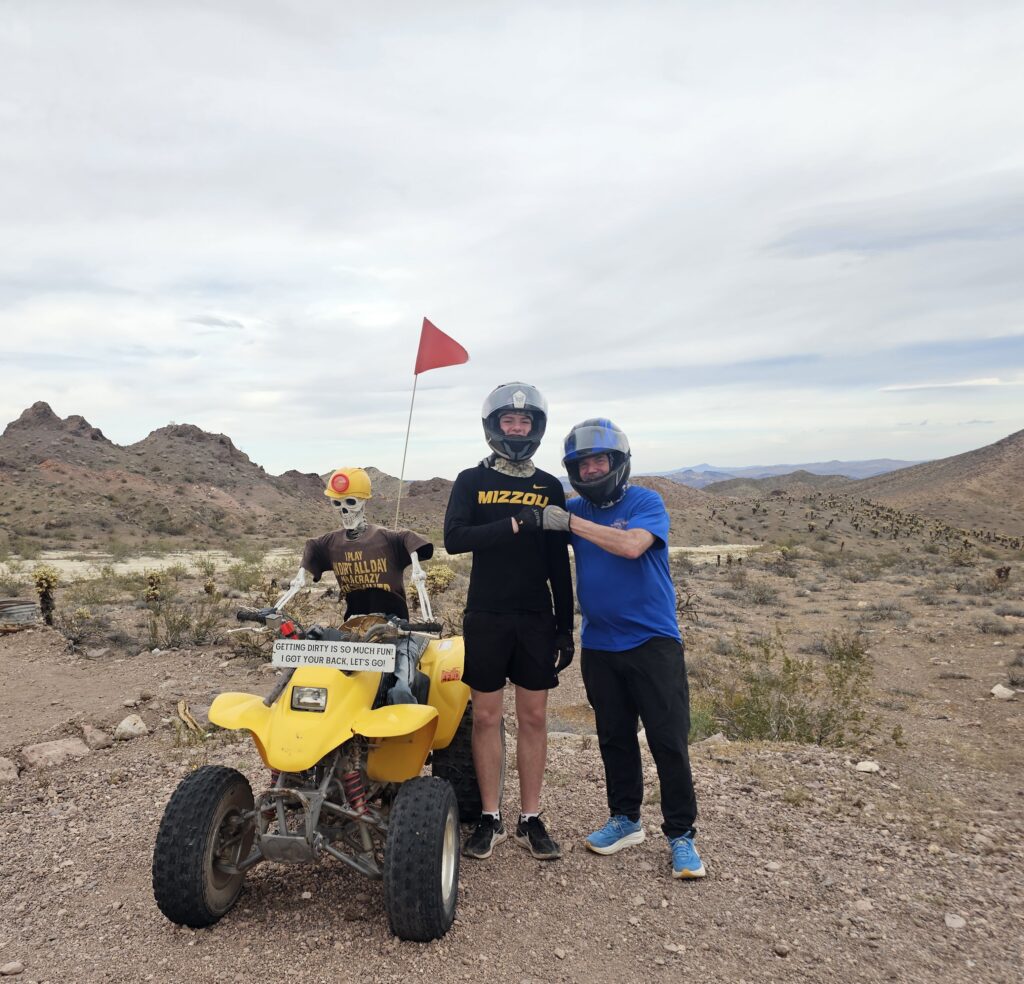 Adventurers in helmets pose by a yellow ATV and skeleton near Nevada’s rocky desert, perfect for Las Vegas Ghost Town ATV tours.