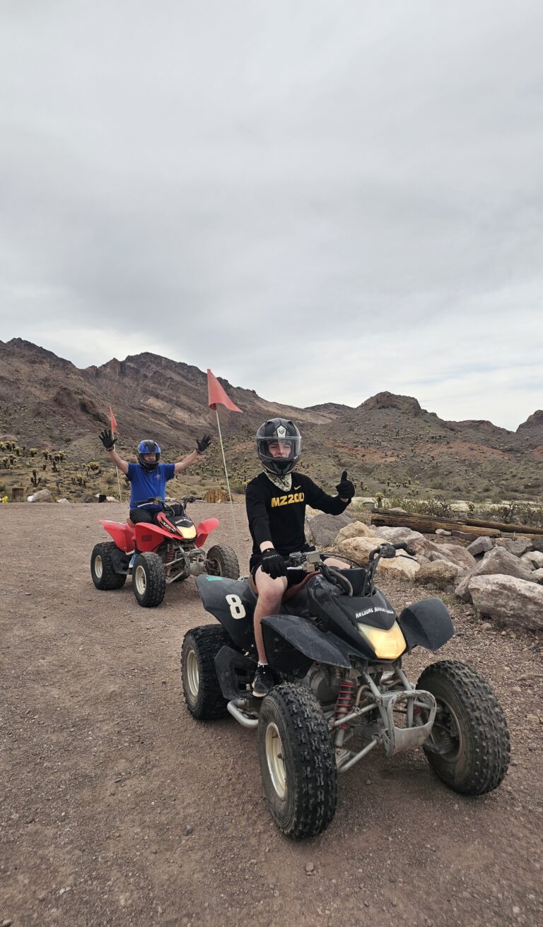 Two people on ATVs giving thumbs-up during a guided Nevada desert ATV tour near Las Vegas, mountains and sky in the background.