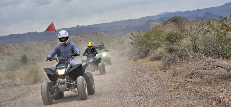 ATV tour near Las Vegas: two helmeted riders with red flags speed along a dusty Nevada desert trail, mountains in the background.